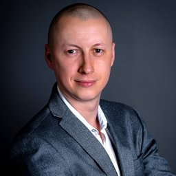 PRESTIGE Nieruchomości Iwona Fryszkowska - Portret mężczyzny w szarej marynarce i białej koszuli, z uśmiechem skierowanym w stronę widza, na ciemnoszarym tle.