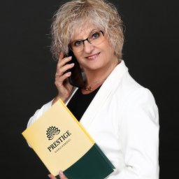 PRESTIGE Nieruchomości Iwona Fryszkowska - Kobieta w białym żakiecie rozmawia przez telefon, trzymając teczkę z logo 'Prestige Nieruchomości' na ciemnym tle. Uśmiechnięta agentka nieruchomości w trakcie rozmowy.