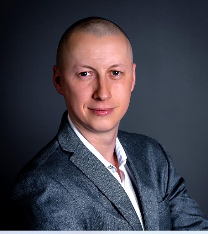 Portret mężczyzny w szarej marynarce i białej koszuli, z uśmiechem skierowanym w stronę widza, na ciemnoszarym tle.