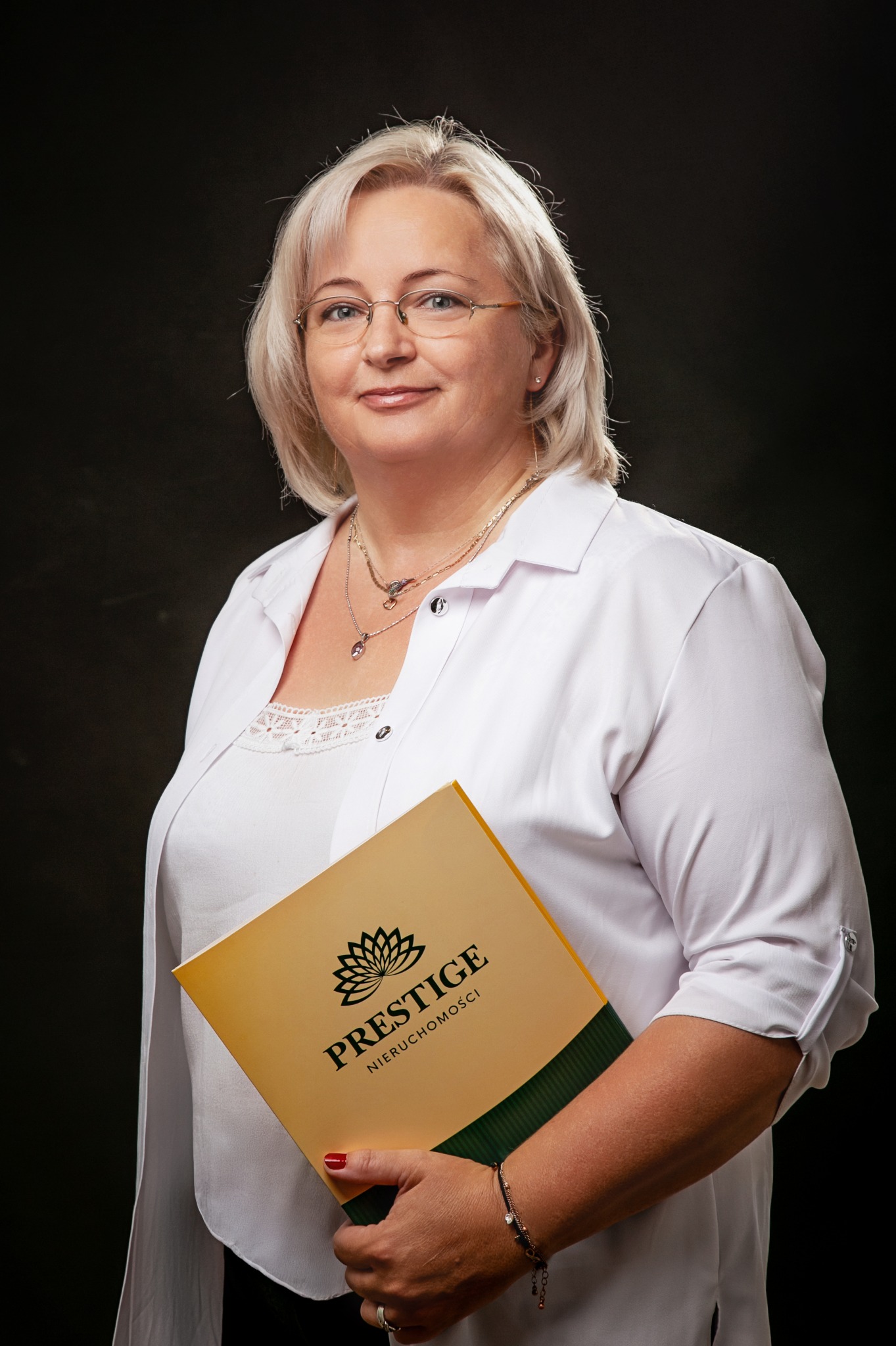 Profesjonalny portret kobiety w okularach, trzymającej folder 'Prestige Nieruchomości' na ciemnym tle. Biała koszula, biżuteria. Uśmiech.