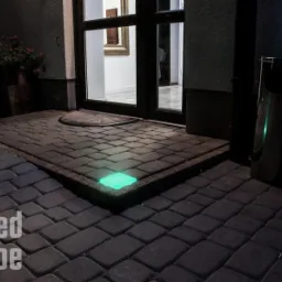 Podświetlona kostka brukowa przy wejściu do budynku, z wbudowanymi, kwadratowymi lampami LED emitującymi zielone światło. Widoczne drzwi wejściowe i donica z kwiatami.