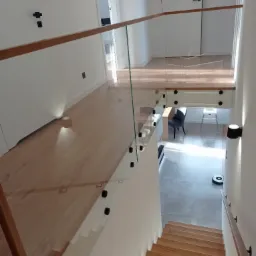 Nowoczesne schody z drewnianymi stopniami i szklaną balustradą mocowaną czarnymi uchwytami, widok z góry na jasne wnętrze z robotem sprzątającym.
