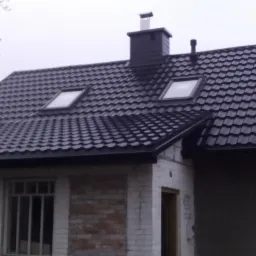 Dom jednorodzinny z czarnym dachem z blachodachówki, dwoma oknami dachowymi i ciemną obróbką blacharską, widoczne rynny, komin i fragment elewacji z cegły oraz tynkowanej