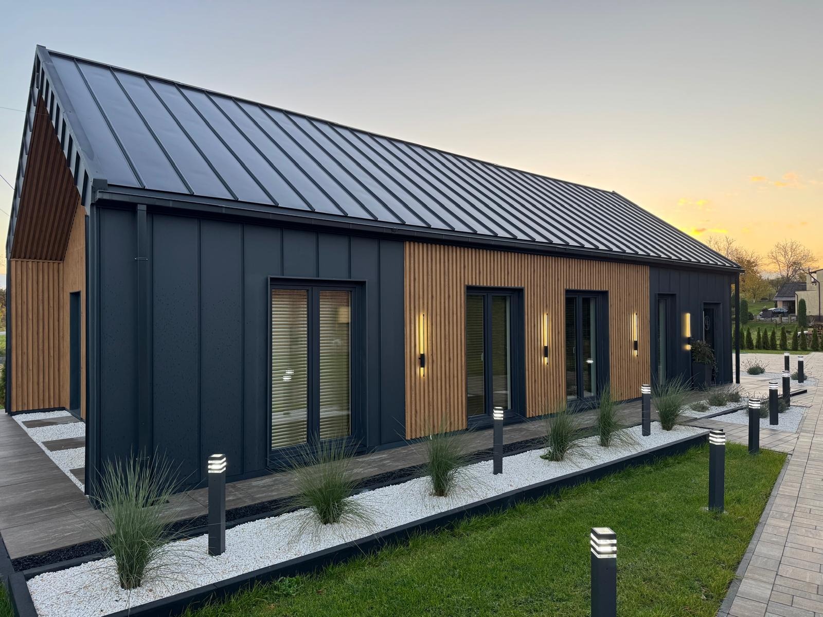 Nowoczesny dom z ciemną blachą na dachu i drewnianą elewacją, oświetlony lampami ogrodowymi. Widok z zewnątrz, podkreślający estetykę i wykończenie.