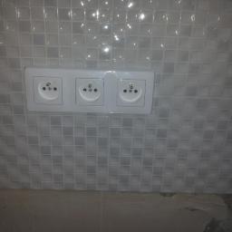 Tomek2014 - Potrójne białe gniazdko elektryczne zamontowane na ścianie wyłożonej błyszczącą, szarą mozaiką kwadratową w łazience lub kuchni.