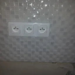 Potrójne białe gniazdko elektryczne zamontowane na ścianie wyłożonej błyszczącą, szarą mozaiką kwadratową w łazience lub kuchni.