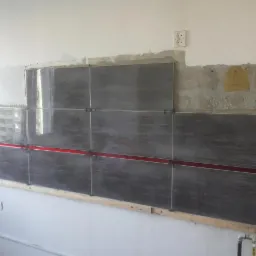 Układanie szarych płytek na ścianie z użyciem poziomicy laserowej i krzyżyków dystansowych. Widoczne przygotowane podłoże i gniazdko elektryczne.