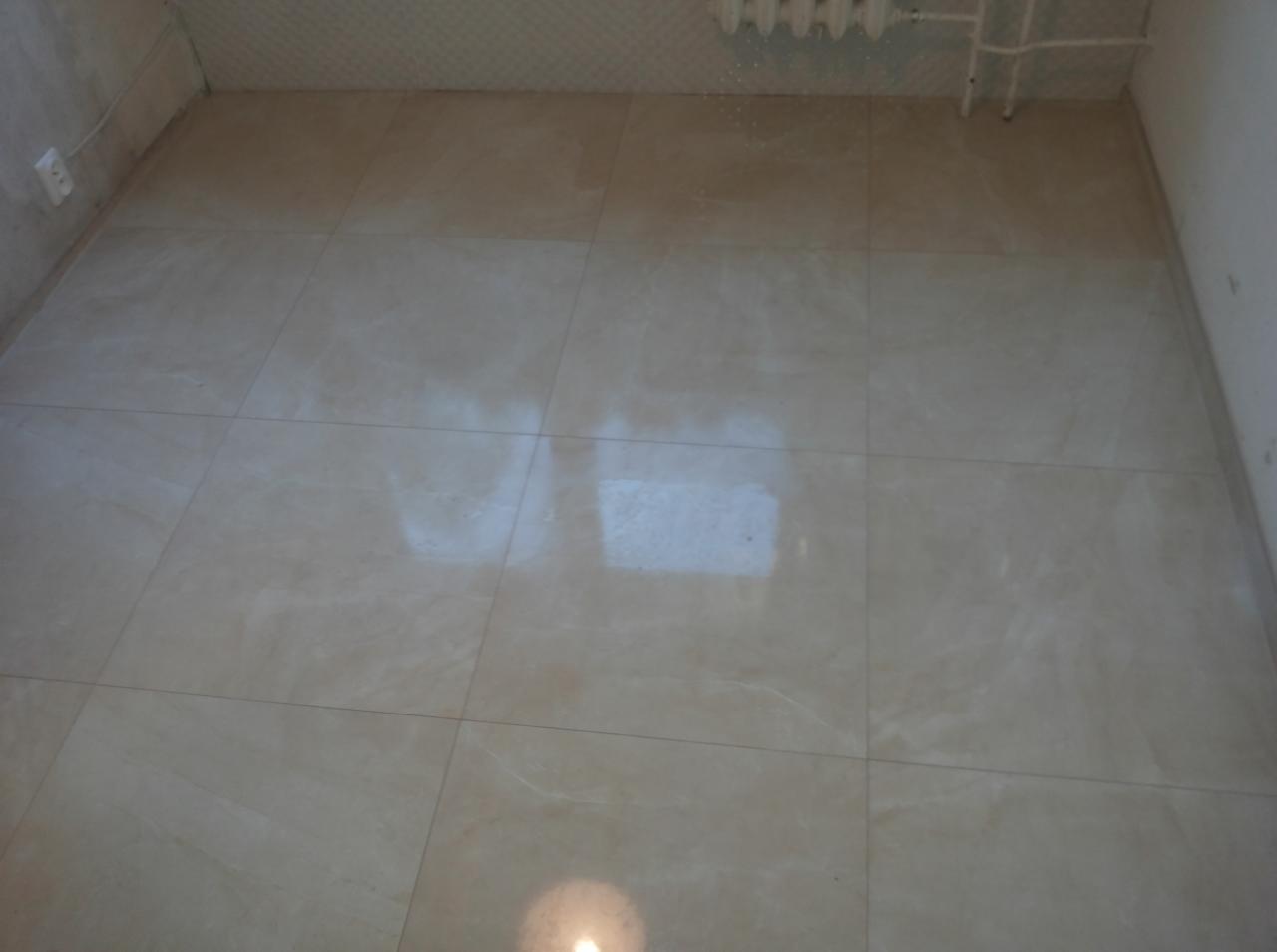 Jasne, błyszczące płytki podłogowe w łazience, ułożone równo, z widocznym grzejnikiem na ścianie i gniazdkiem elektrycznym. Podłoga odbija światło, tworząc jasne refleksy.