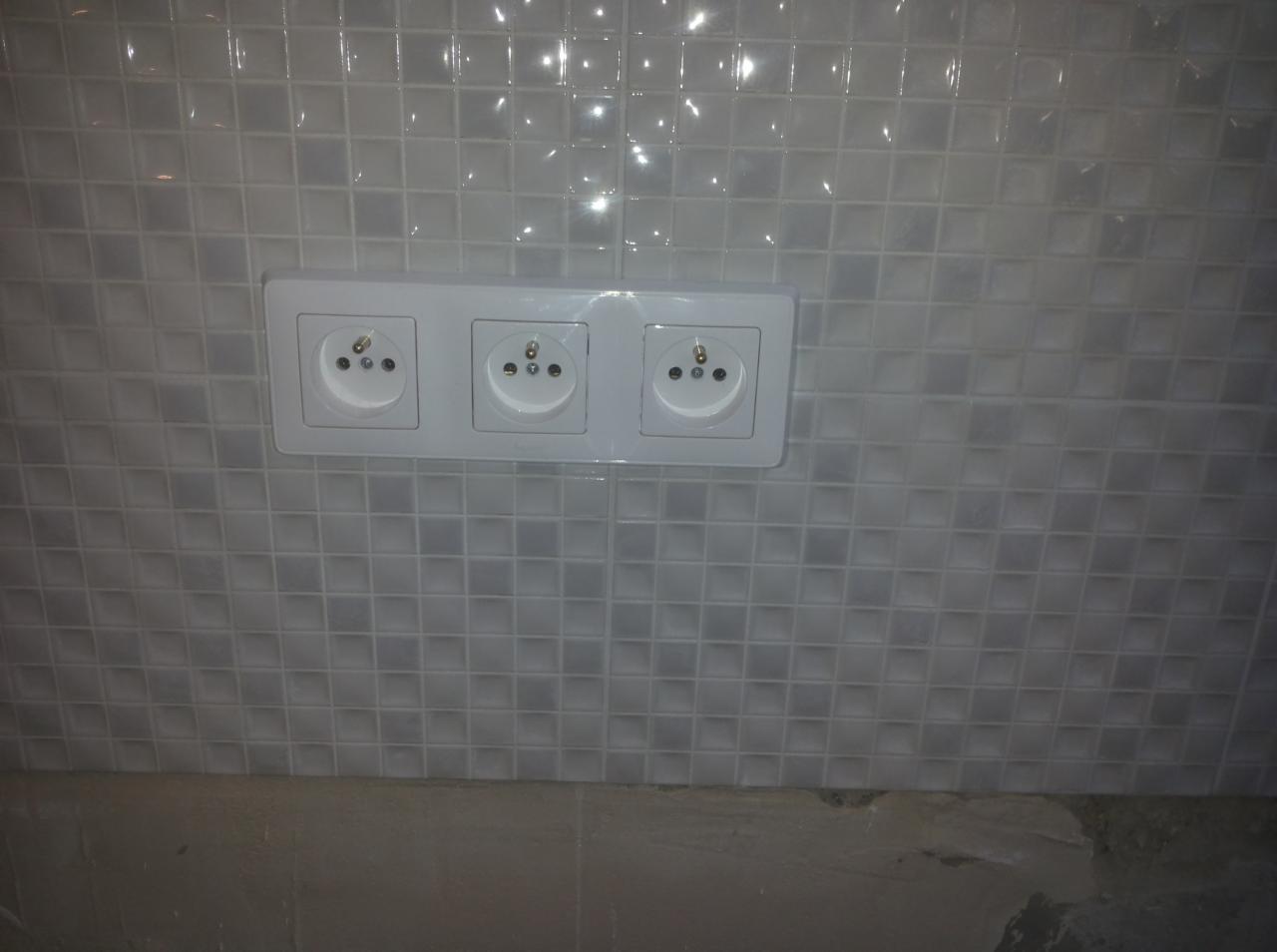 Potrójne białe gniazdko elektryczne zamontowane na ścianie wyłożonej błyszczącą, szarą mozaiką kwadratową w łazience lub kuchni.