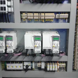 Szara szafa sterownicza z zamontowanymi białymi falownikami ABB i licznymi złączkami elektrycznymi. Widoczne przewody i kable. Detal instalacji elektrycznej w przemysłowej szafie.