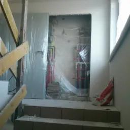 Remont klatki schodowej w trakcie: schody z płytek, zabezpieczone rury instalacji za folią, drabina. Prace wykończeniowe w budynku.