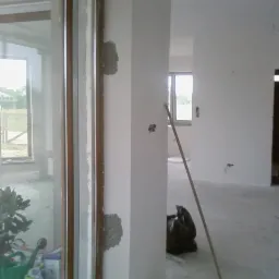 Wnętrze w trakcie prac wykończeniowych: gładzie na ścianach, okno z widokiem na zewnątrz, podłoga przygotowana do dalszych prac, widoczna roślina w doniczce.