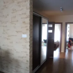 Wykończenie ściany imitacją kamienia obok ciemnej szafy przesuwnej. Widok na parkiet i drzwi w głębi mieszkania. Dekoracyjny detal w nowoczesnym wnętrzu.