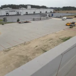 Rozległy, betonowy plac manewrowy przed budynkiem magazynowym z rampami załadunkowymi, z widocznymi pojazdami budowlanymi, stertą kostki brukowej oraz wykopem w tle, sugerujący prace ziemne...