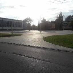 Nowo wyłożony plac z kostki brukowej w kształcie łuku, połączony z asfaltową drogą, otoczony budynkami i drzewami w słoneczny dzień.