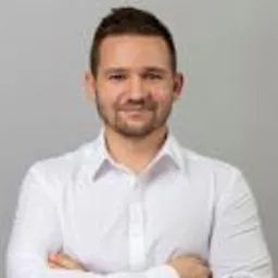 Portret mężczyzny w białej koszuli na jasnoszarym tle, uśmiechnięty, z krótką brodą i zaczesanymi włosami.