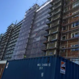 Wielopiętrowy budynek w trakcie remontu, osłonięty siatką ochronną, rusztowania na całej elewacji. Kontener HKL na pierwszym planie, błękitne niebo w tle.