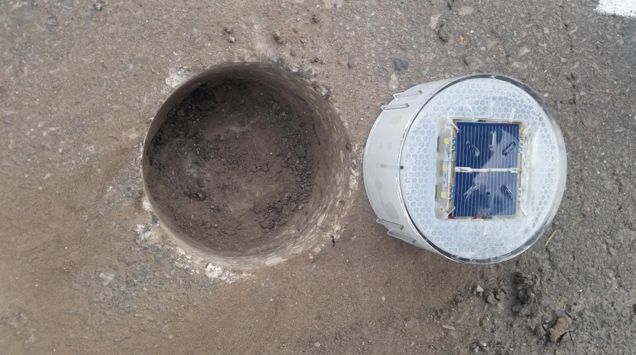 Przygotowany otwór w asfalcie obok leżącego, okrągłego, solarnego punktowego elementu odblaskowego drogowego w obudowie z tworzywa.