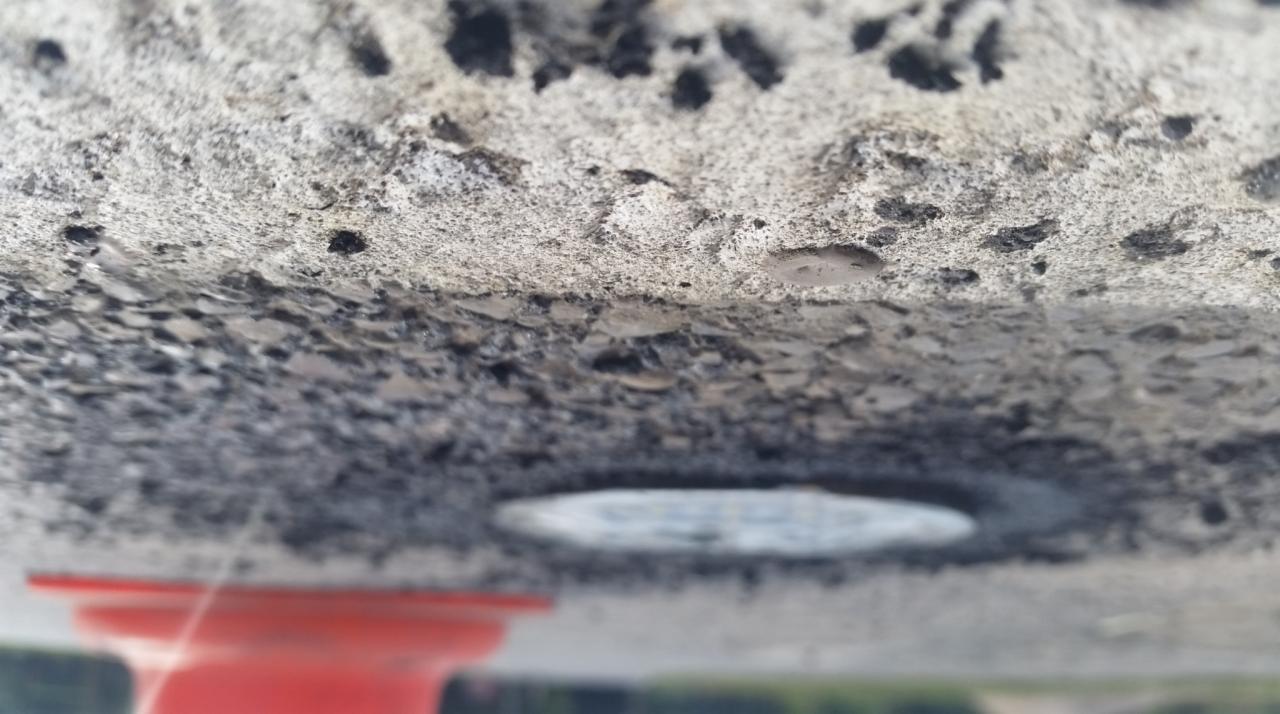 Betonowa konstrukcja z wbudowanym okrągłym elementem, prawdopodobnie oprawą oświetleniową, widziana z bliska od dołu, z rozmytym czerwonym elementem i zielenią w tle.