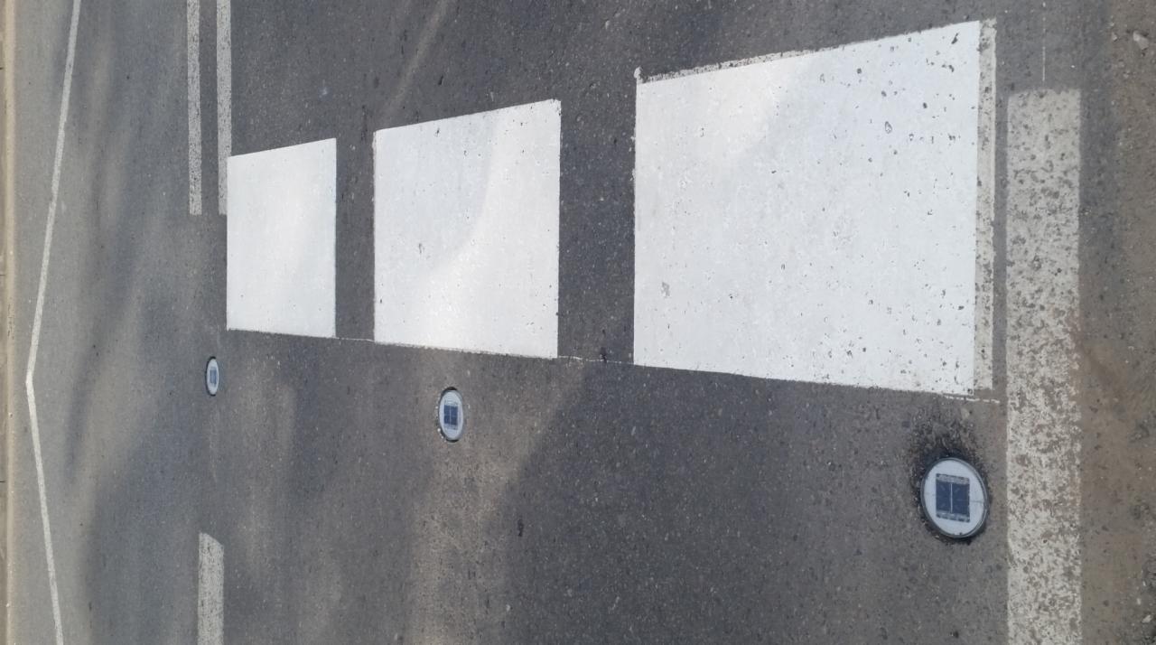 Fragment jezdni z wymalowanym przejściem dla pieszych i wbudowanymi w asfalt lampami solarnymi.
