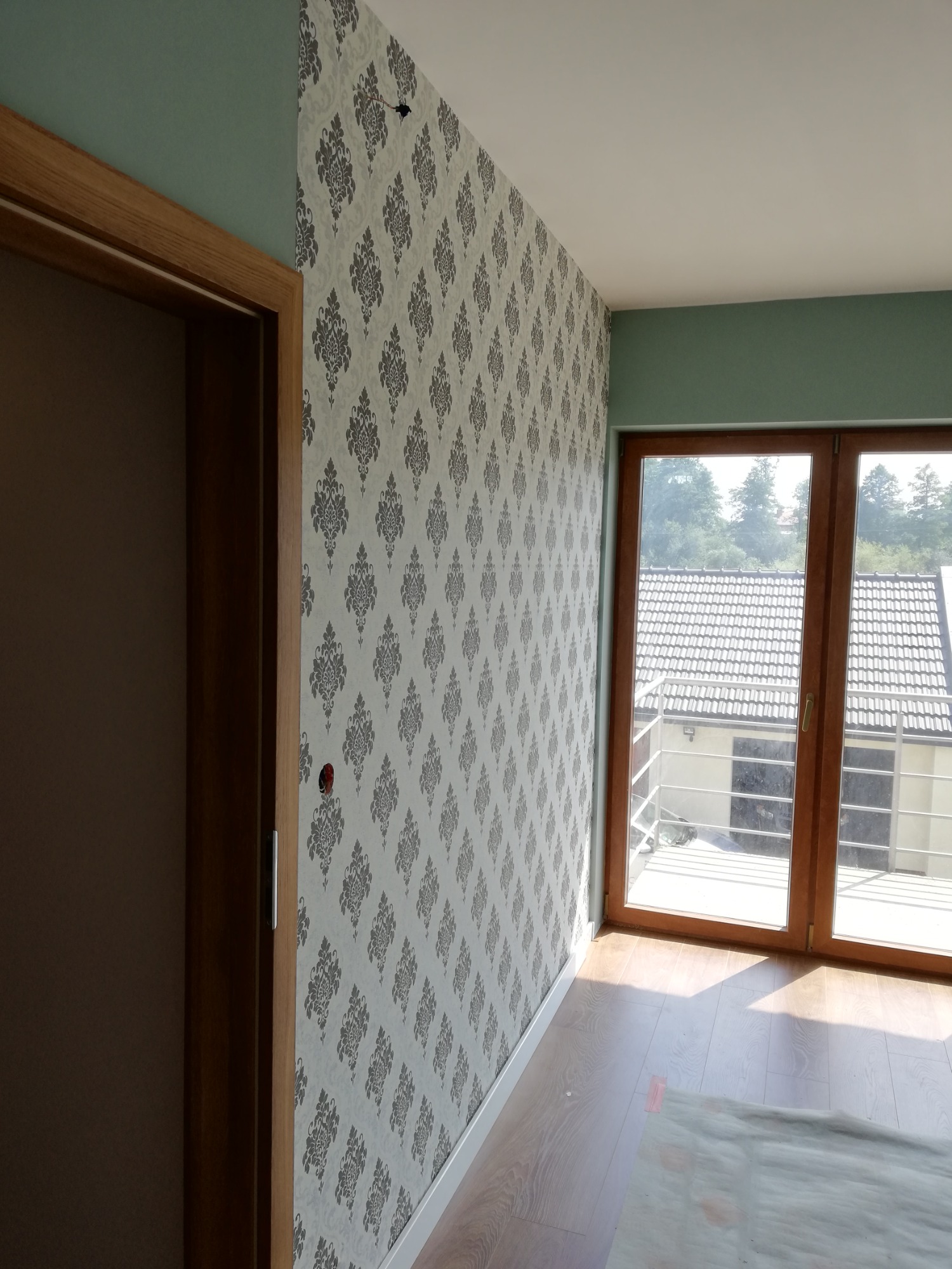 Pokój po remoncie z tapetą w szaro-białe wzory, drewnianą stolarką okienną i drzwiową, oraz fragmentem podłogi zabezpieczonej folią malarską.