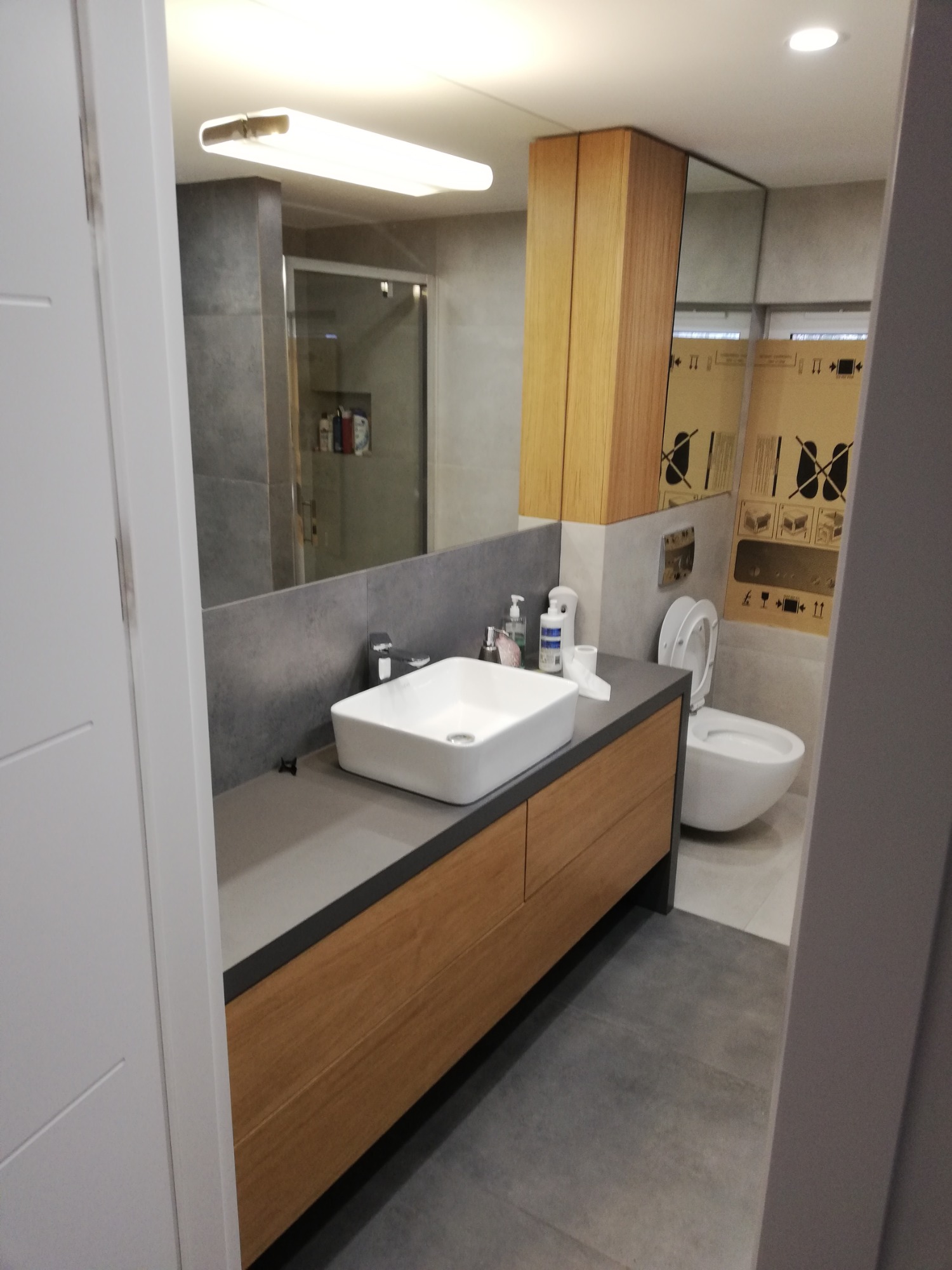 Nowoczesna łazienka z drewnianą szafką pod umywalką, szarą glazurą i widoczną toaletą w tle, częściowo zasłoniętą kartonem ochronnym.