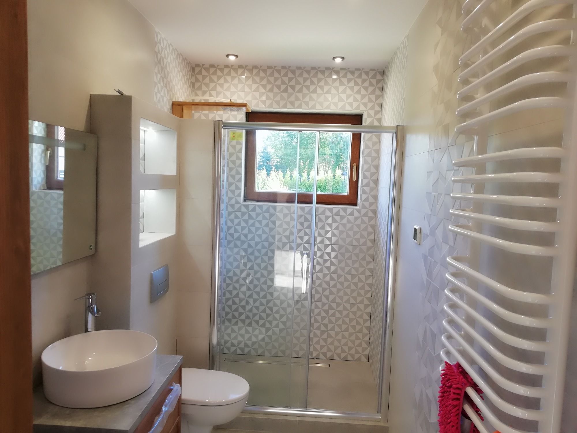 Nowoczesna łazienka z kabiną prysznicową z szybą, umywalką nablatową na szafce z szarą płytą, grzejnikiem drabinkowym z różowym ręcznikiem i geometryczną mozaiką na ścianach.