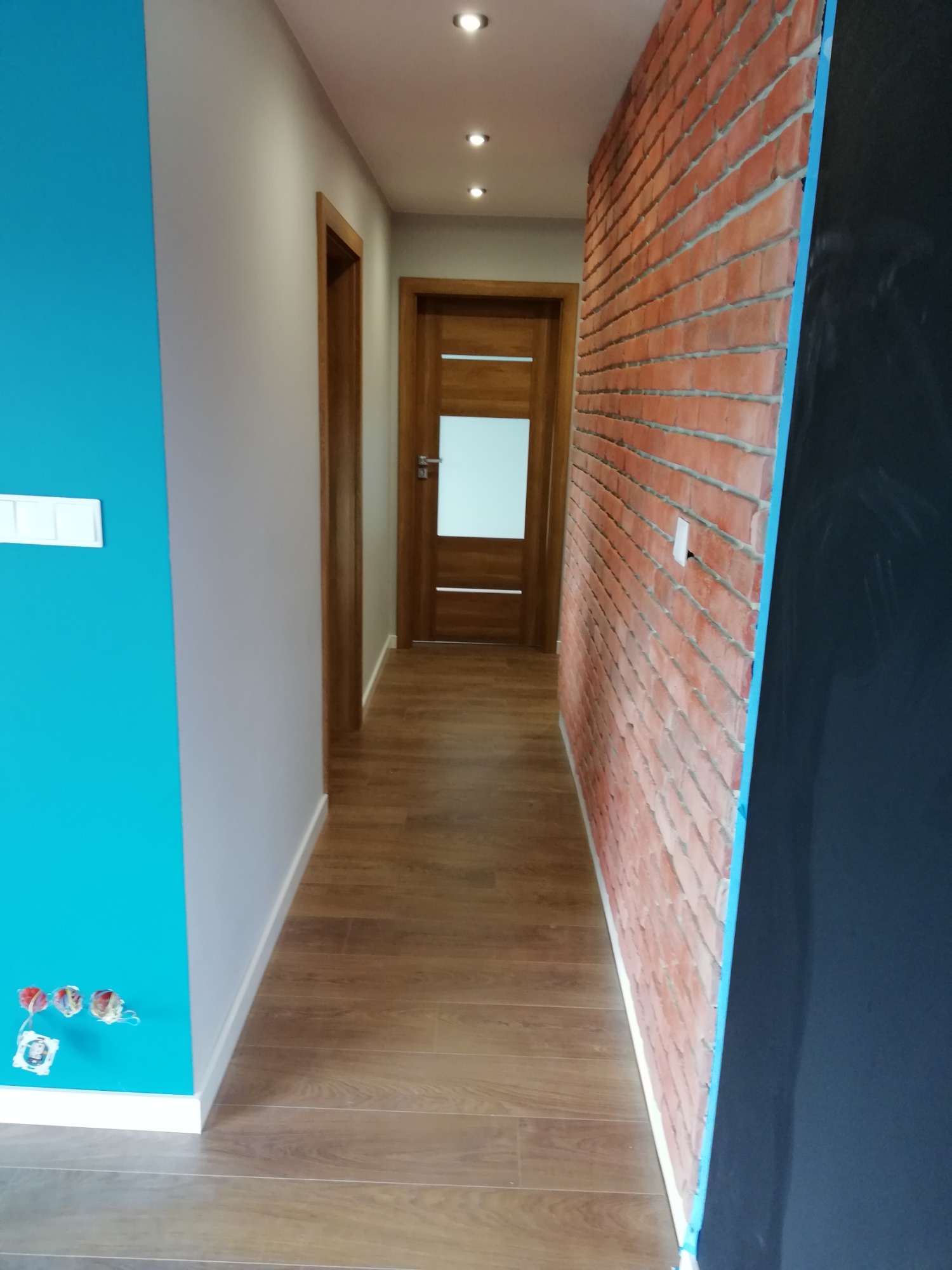 Wnętrze korytarza po odświeżeniu: turkusowa ściana z wystającymi przewodami elektrycznymi, szara ściana, imitacja cegły na ścianie, drewnopodobne panele na podłodze, drzwi z przeszkleniem w głębi