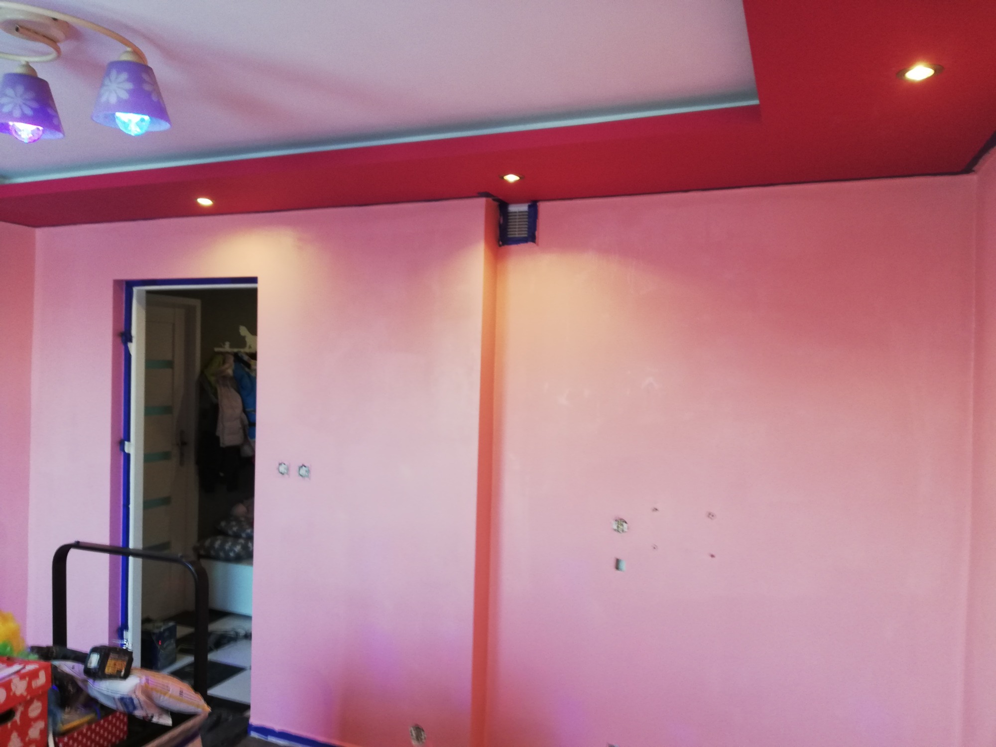 Pokój pomalowany na różowo z czerwonym sufitem, widoczne ślady po naprawach na ścianie, lampa sufitowa z fioletowymi kloszami i otwarte drzwi do garderoby