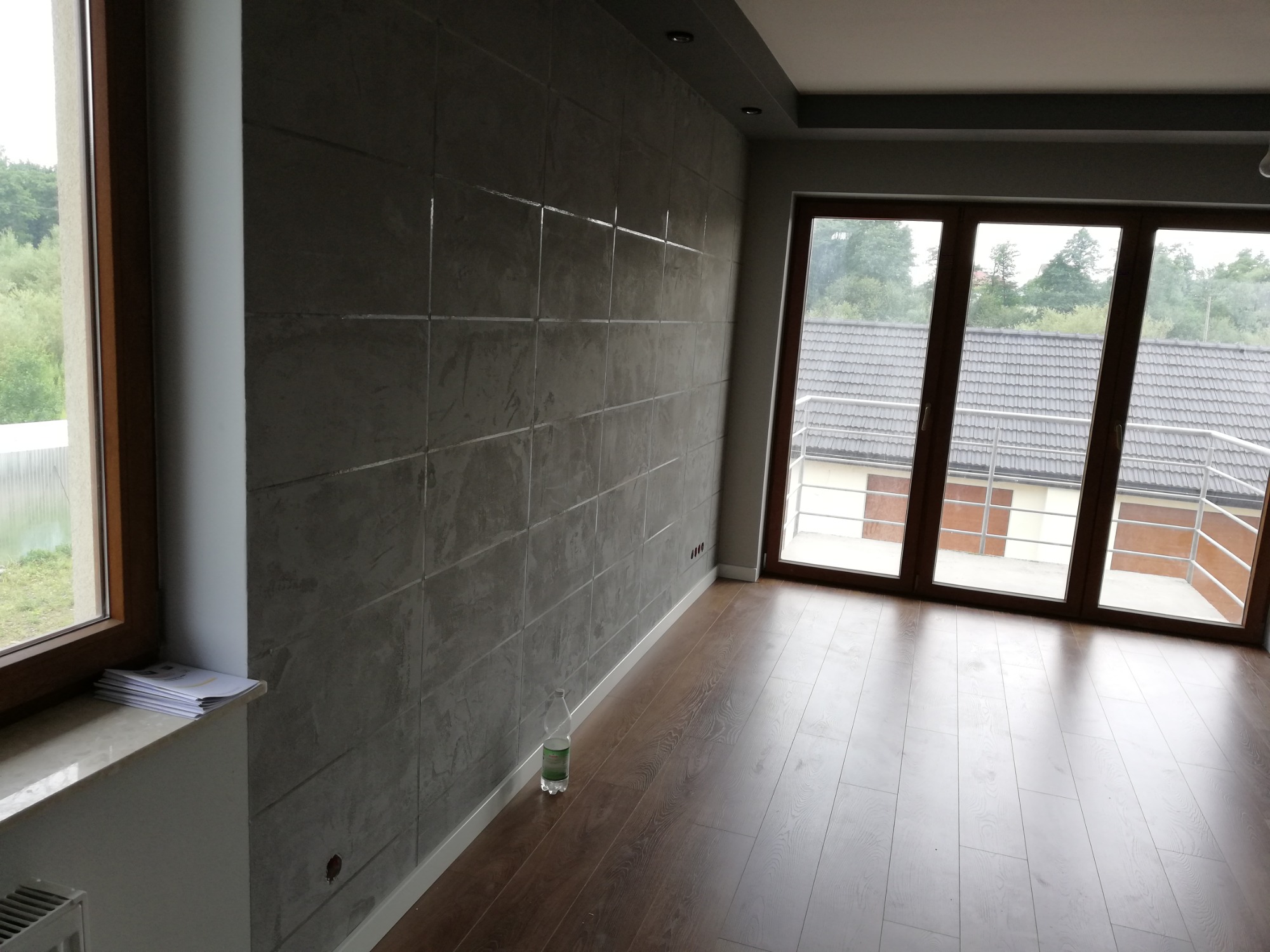 Wnętrze pokoju z panelami podłogowymi w odcieniach brązu, ściana wykończona szarymi płytami imitującymi beton, duże okna z widokiem na ogród i sąsiedni dom.