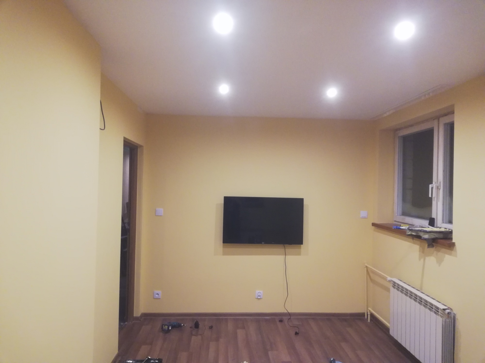 Pokój z panelami podłogowymi i żółtymi ścianami, na ścianie wisi telewizor, widoczne narzędzia na podłodze i grzejnik pod oknem.