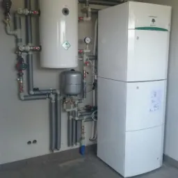 Nowoczesna instalacja grzewcza z zbiornikiem i białą jednostką grzewczą, rurami i manometrem na szarej ścianie w surowym pomieszczeniu z szarą podłogą.