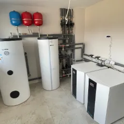 Kotłownia z dwoma zbiornikami akumulacyjnymi, naczyniami wzbiorczymi i dwoma jednostkami grzewczymi, rury w izolacji, jasne płytki podłogowe.