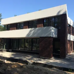 Nowoczesny dom z cegły klinkierowej z dużymi oknami i białymi elementami architektonicznymi, widok z zewnątrz, teren w trakcie prac wykończeniowych.