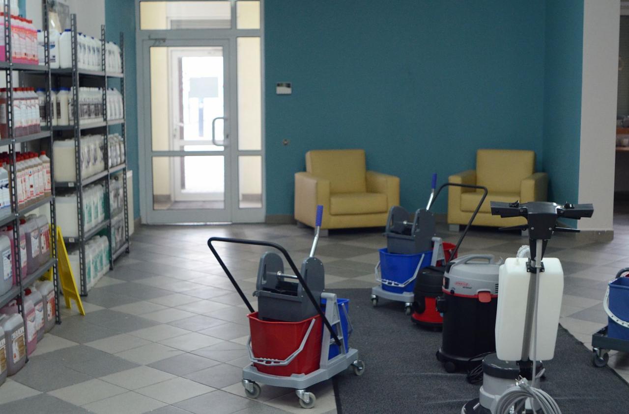 Wnętrze z wózkami do sprzątania, odkurzaczem i regałami pełnymi środków czystości. W tle dwa żółte fotele i przeszklone drzwi wejściowe.
