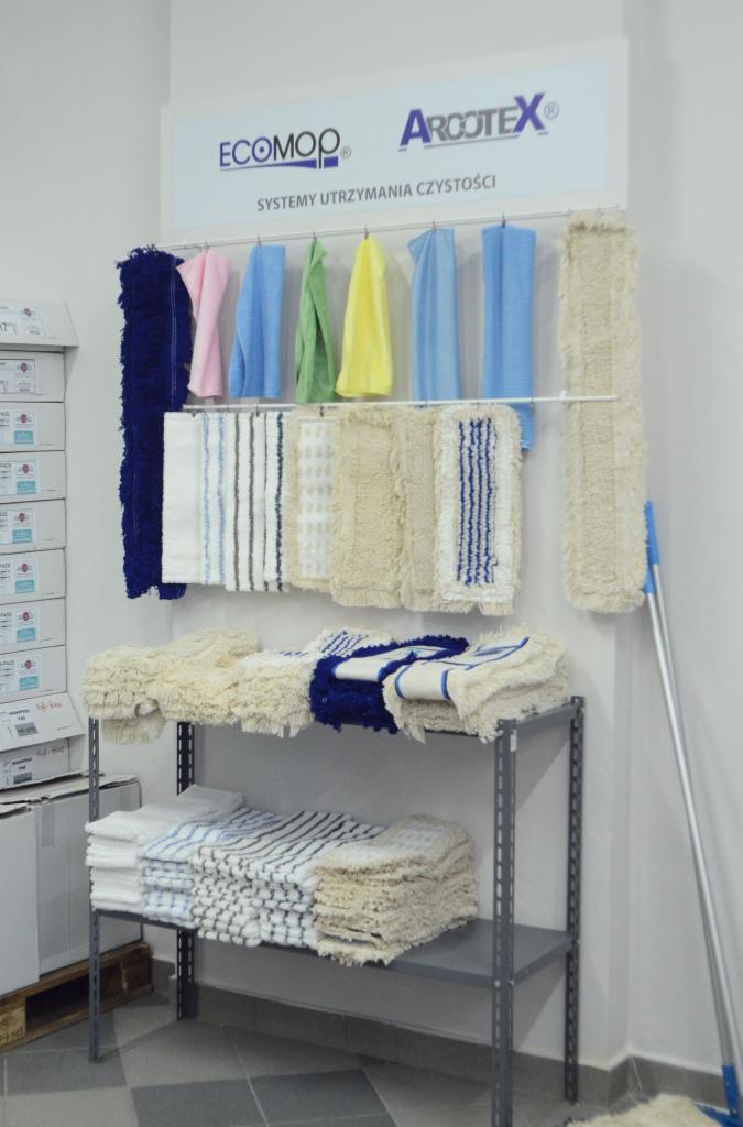 Ekspozycja ścierek i mopów firmy ARCOTEX na metalowym regale w sklepie, obok widoczne kartony z produktami.