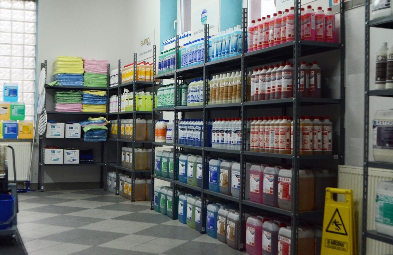 Półki regałowe w sklepie ARCORA SHOP Łódź, wypełnione różnorodnymi butelkami, kanistrami i pojemnikami z płynami oraz stertami ścierek i mopów, widoczny znak ostrzegawczy o śliskiej podłodze.