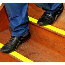 Stopa w czarnym bucie sportowym na drewnianym schodku z żółtą taśmą antypoślizgową Safety Walk 3M, widok z góry pod kątem.