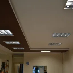 Dwukolorowy sufit z brązowym pasem i wpuszczonymi lampami rastrowymi, widok na wnętrze pomieszczenia z elementami wyposażenia.