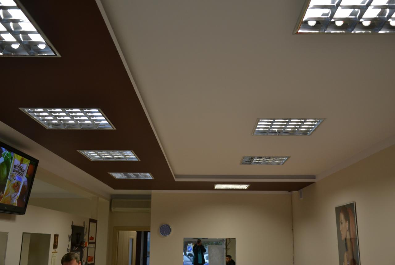 Dwukolorowy sufit z brązowym pasem i wpuszczonymi lampami rastrowymi, widok na wnętrze pomieszczenia z elementami wyposażenia.