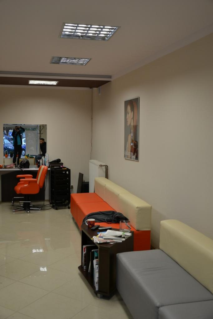 Wnętrze salonu fryzjerskiego z pomarańczowym fotelem, stanowiskiem pracy z lustrem, sofą w odcieniach beżu, pomarańczy i szarości oraz stolikiem z gazetami i kubkiem.