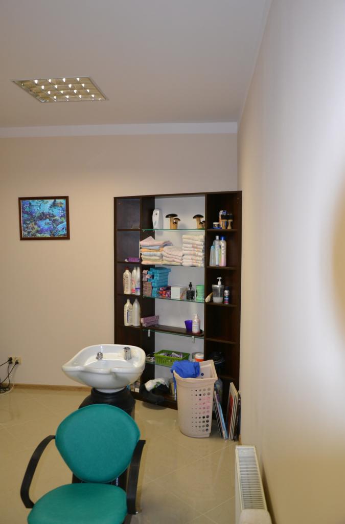 Wnętrze salonu fryzjerskiego z umywalką fryzjerską, krzesłem w kolorze turkusowym i regałem z kosmetykami oraz ręcznikami, oświetlone lampą sufitową.