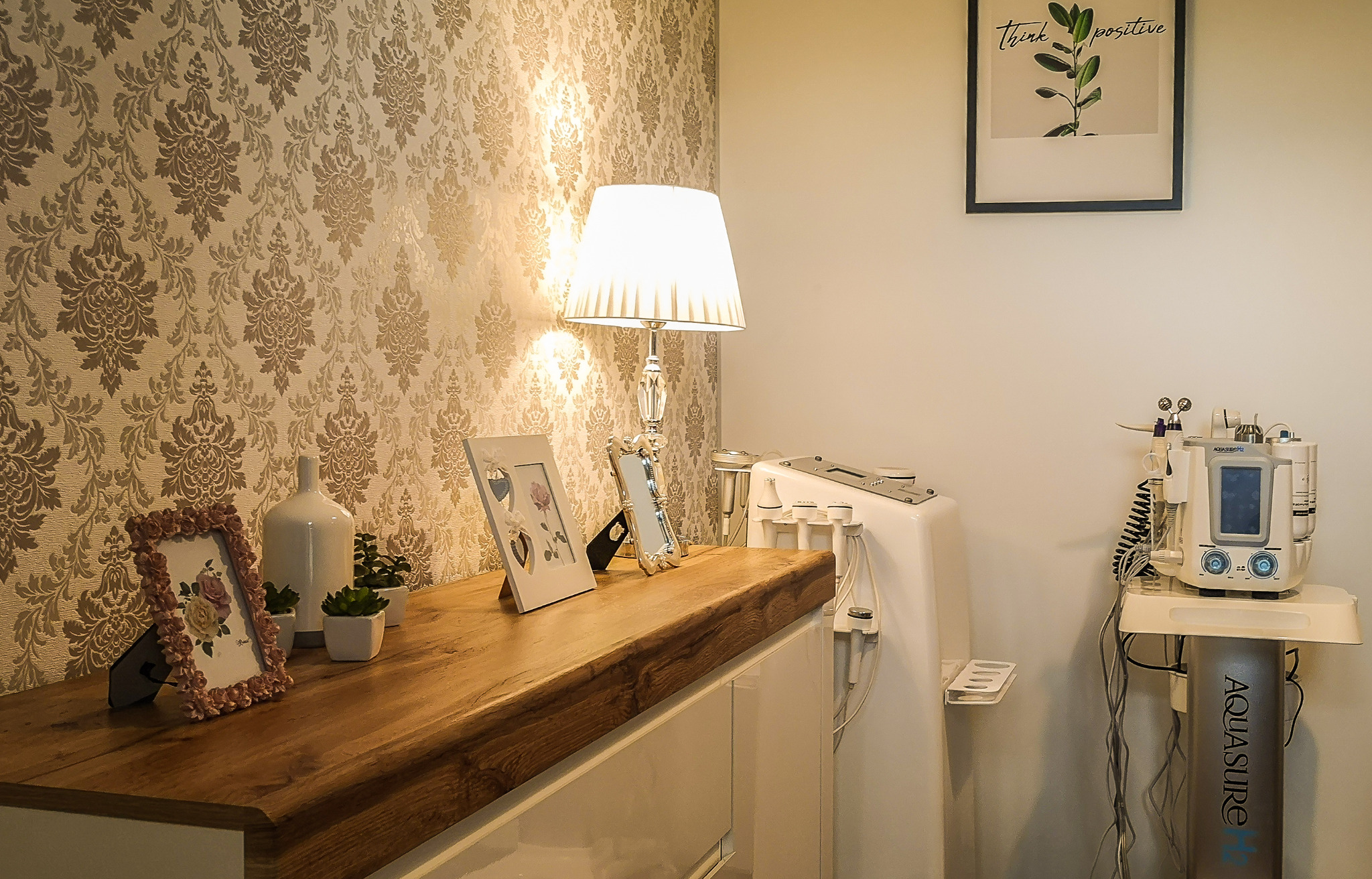 Gabinet kosmetyczny: widok na drewniany blat z dekoracjami (ramki, wazon, sukulenty) i profesjonalne urządzenie kosmetyczne Aquasure H2 w tle, ozdobna tapeta w kwiaty.