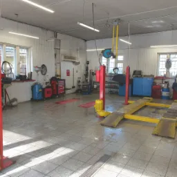 Wnętrze warsztatu samochodowego z dwoma podnośnikami kolumnowymi w kolorze czerwonym i żółtym, narzędziami i wyposażeniem warsztatowym pod ścianami, oraz oświetleniem jarzeniowym.