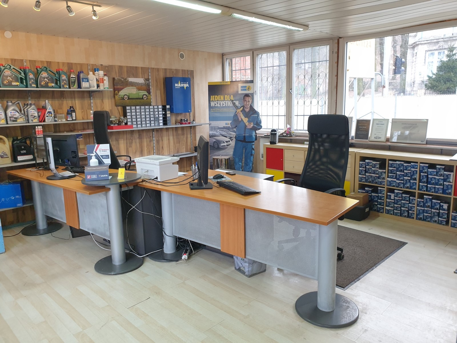 Wnętrze biura warsztatu samochodowego z biurkiem, komputerami, olejami silnikowymi na półkach, plakatem reklamowym Bilstein i certyfikatami na szafce przy oknie.