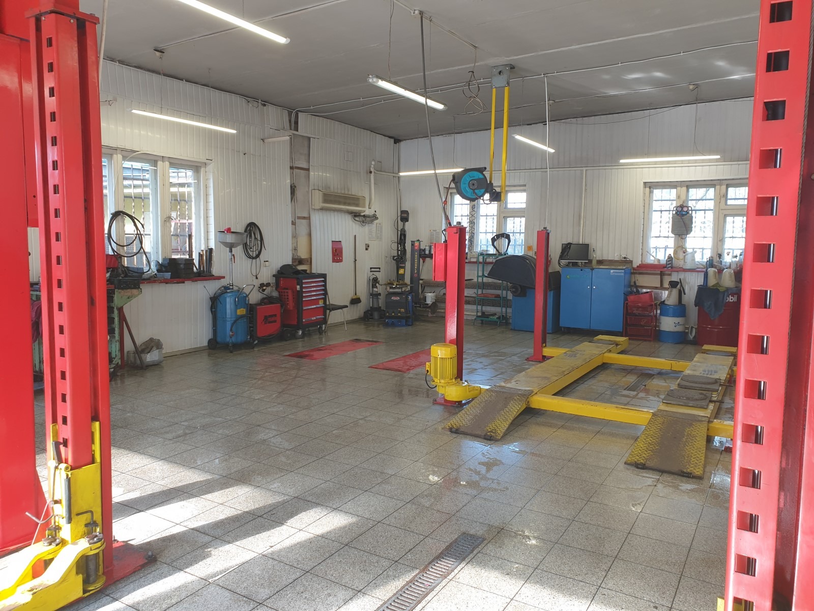 Wnętrze warsztatu samochodowego z dwoma podnośnikami kolumnowymi w kolorze czerwonym i żółtym, narzędziami i wyposażeniem warsztatowym pod ścianami, oraz oświetleniem jarzeniowym.