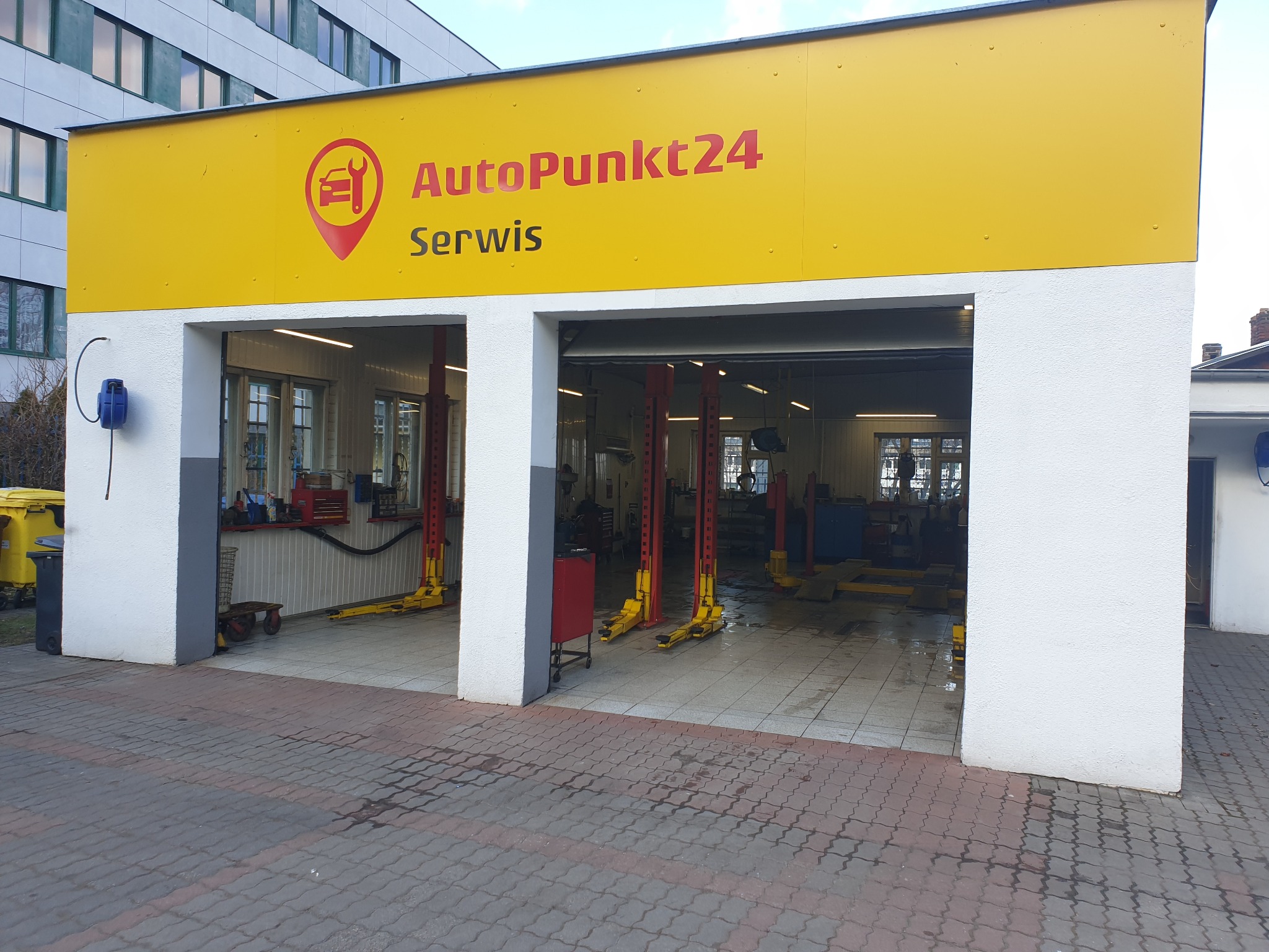 Widok z zewnątrz na warsztat samochodowy AutoPunkt24 z trzema stanowiskami, każdy wyposażony w podnośnik kolumnowy, widoczne narzędzia i wyposażenie wewnątrz.