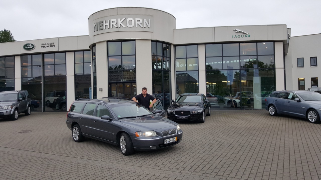 Szary samochód Volvo kombi z otwartymi drzwiami i mężczyzną opierającym się o dach, na tle salonu samochodowego z logo Jaguar i Land Rover.