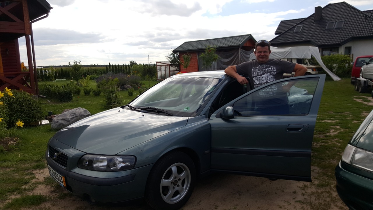 Zielony samochód osobowy marki Volvo z otwartymi drzwiami, obok stoi mężczyzna w szarej koszulce z nadrukiem, w tle dom z czerwonego drewna i zabudowania gospodarcze.
