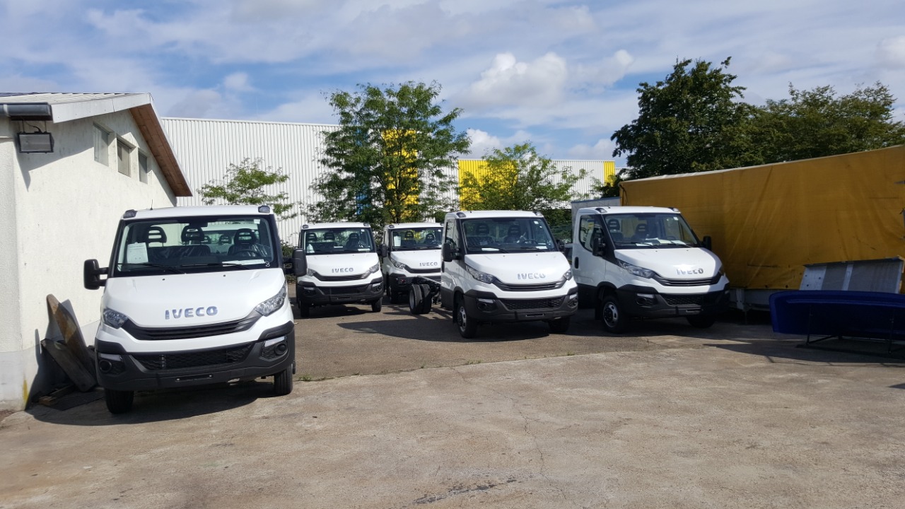 Rząd białych samochodów dostawczych Iveco na parkingu, jeden bez zabudowy, w tle budynek z żółtą ścianą i drzewa.